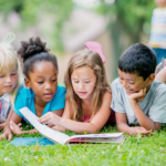 four children of different ethnicities sitting on the grass reading children's books together