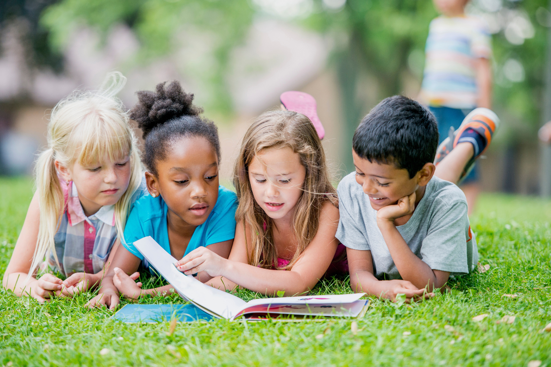 four children of different ethnicities sitting on the grass reading children's books together